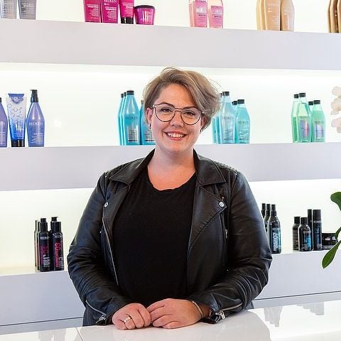 Debbie, eigenaresse van Marlee Hair, lachend in haar salon voor de schappen met Redken-producten